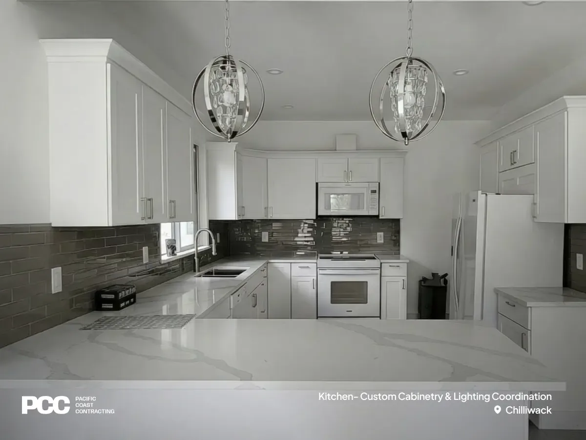 A straight-on view of a modern white kitchen remodel by Pacific Coast Contracting in Chilliwack. The bright space highlights custom white shaker cabinetry, striking spherical lighting over a quartz peninsula, stainless steel appliances, and a sleek grey tiled backsplash by Pacific Coast Contracting.